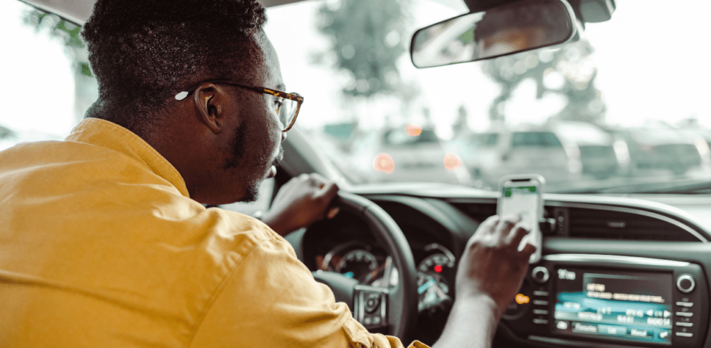 Man in yellow shirt using GPS on smartphone while driving in traffic, focusing on the car’s dashboard