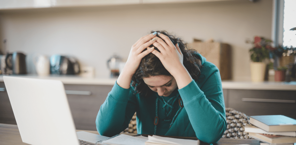 trauma from a vehicle accident in Las Vegas Young woman in a kitchen with her head in her hands, appearing stressed, with a laptop and notebooks on the table