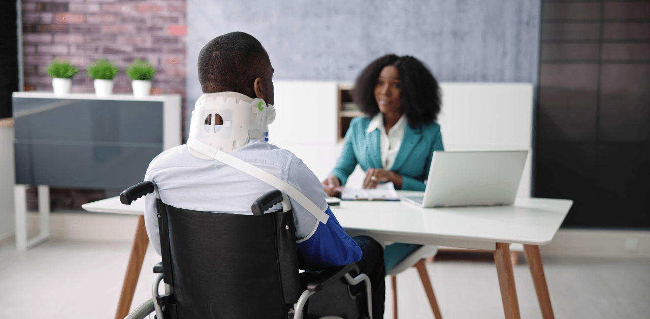 A person in a wheelchair wearing a neck brace, sitting across a desk from a business professional during a meeting