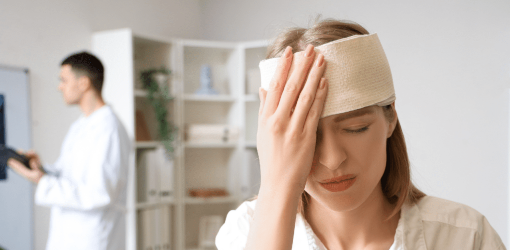 Woman with a bandaged head holds her forehead in pain while a doctor in the background examines medical records in a clinic