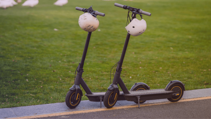 Two electric scooters with helmets hanging from the handlebars parked beside a grassy area