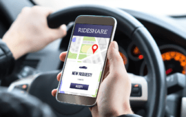 Driver holding a smartphone displaying a rideshare app with a new ride request while seated behind the wheel of a car