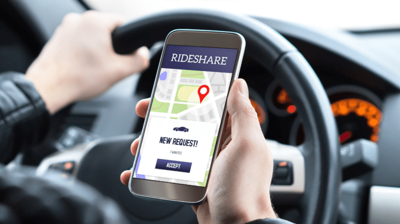 Driver holding a smartphone displaying a rideshare app with a new ride request while seated behind the wheel of a car