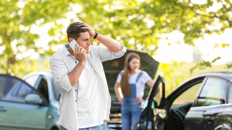 A man on the phone looking distressed after a car accident, with a woman standing near another vehicle in the background