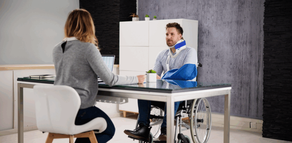 Man in a wheelchair with a neck brace and arm sling speaking to a woman across a desk in an office setting
