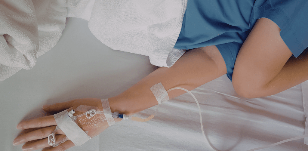 Patient lying in a hospital bed with an intravenous IV line attached to their hand beneath a blanket